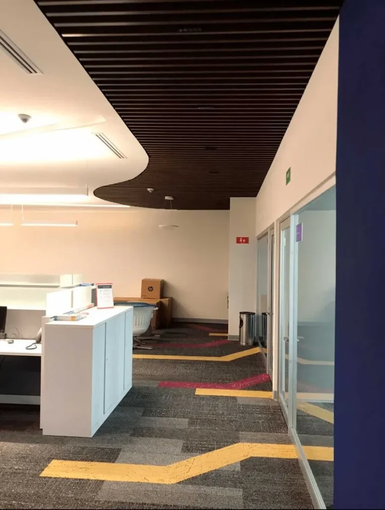 Interior de oficina mostrando un techo con listones de madera oscura y una alfombra gris con patrones amarillos y rojos