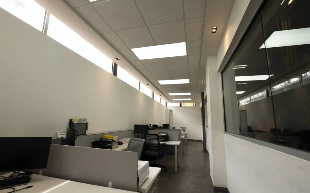 Pasillo de oficina con ventanales superiores que permiten entrada de luz natural sobre escritorios blancos y sillas negras