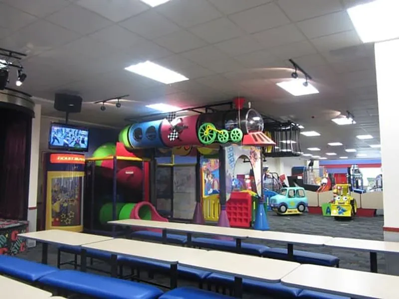 Interior con laberinto de tubos de colores y mesas largas con bancas azules bajo un techo de oficina con luces LED