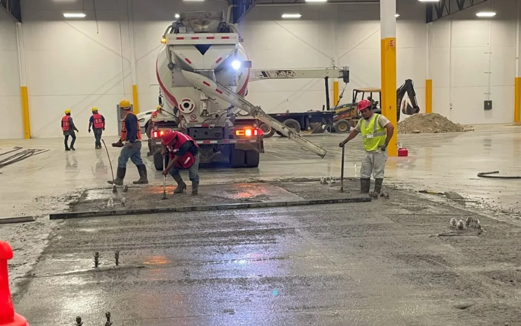 Trabajadores nivelando concreto fresco vertido por camión revolvedor en el piso reforzado de una nave industrial.