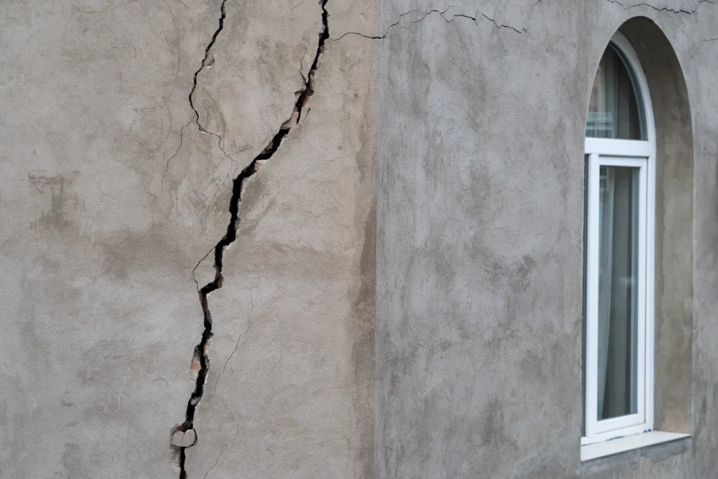 Fotografía de una construcción de paredes color gris con una ventana blanca que tiene fisuras y grietas en un costado.