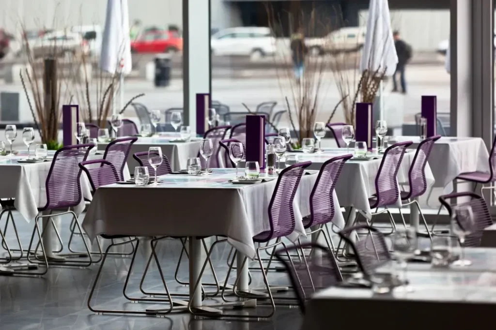 Interior moderno de restaurante con mesas blancas, sillas moradas y grandes ventanales.